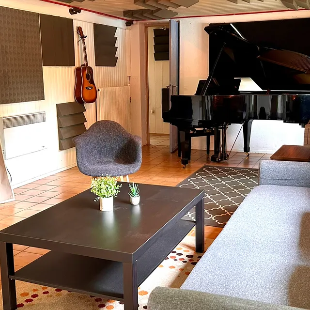 Vue large du salon acoustique : piano à queue, canapé gris, table basse et cuisine ouverte, plafond équipé de pièges à basses